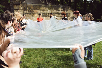 A group of people hold a white sheet between them on a lawn.