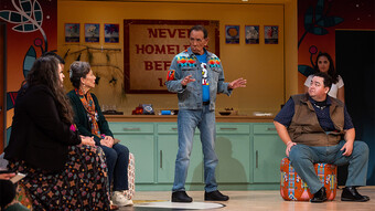 An actor in a denim jacket stands center, speaking to a group of seated actors.