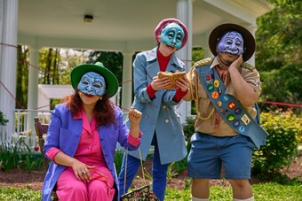 Three actors in brightly colored clothing and masks perform outside.