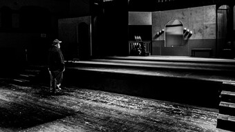 A man stands with his back to the camera and stares at an empty set on stage.
