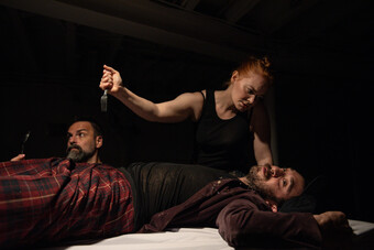 An actors holds a fork over an actor lying on a table, as if preparing to stab him.