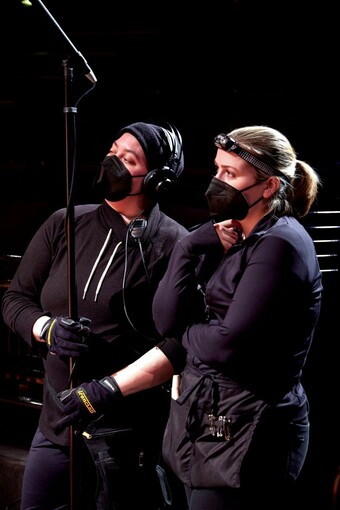 Two people in stage blacks wearing masks stand backstage.