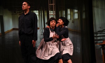 two actors in aprons sit on stage next to an actor standing with his back to them.