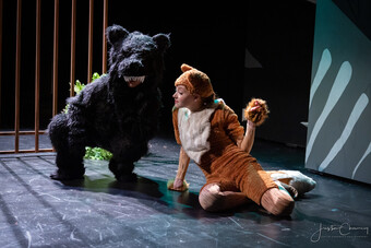 Two actors wearing fuzzy animal costumes sit on stage.