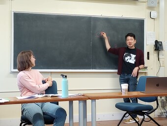 A writer stands in front of a blank chalkboard in the front of a classroom.