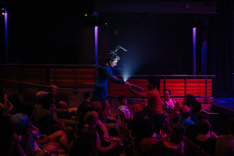 A performer wearing a helmet walks into a dimly lit audience and holds hands with an audience member.