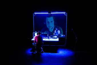 A man using computers and displaying his face on a large screen. 
