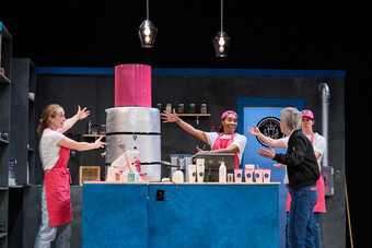 Three people dressed in pink aprons and hats present a pink vat to someone dressed as a manager on stage. 