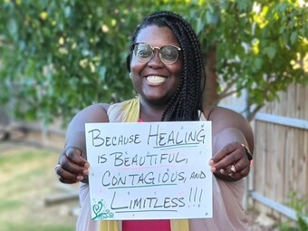 A black woman holding a sign that reads "because healing is beautiful, contagious, and limitless."