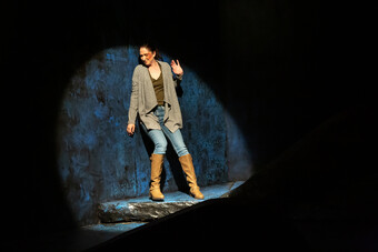 A woman in jeans and a grey cardigan flinching in a spotlight. 
