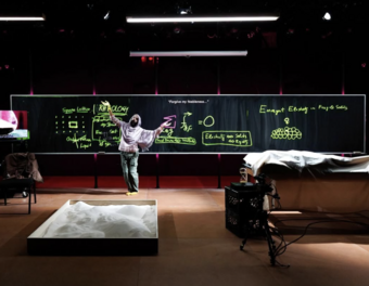 A person in a shawl standing in front of a black board with neon green writing on it.