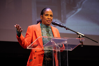 A woman in an orange blazer giving a speech at a clear podium.