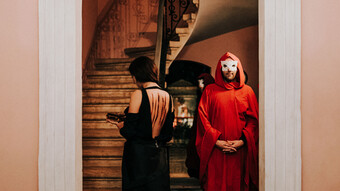 An actor in a red robe and white mask in a stairway with another actor in a black backless dress.