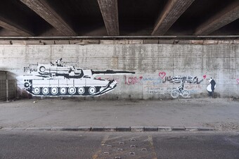A wall painted with the image of a tank pointing its gun at a person on a bike.