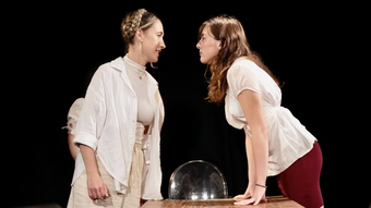 Two women on stage standing up and looking at each other over a table.