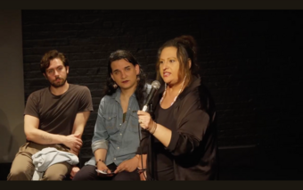 A person on stage speaking into a microphone while two people look on.
