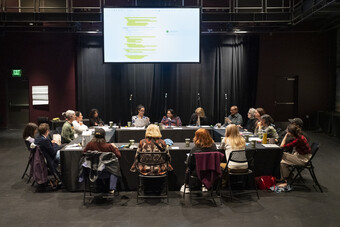 A group of people sit around a table in a black box theatre.