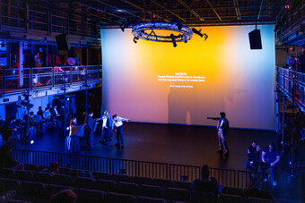 A group of performers stand onstage with supertitles in the background.