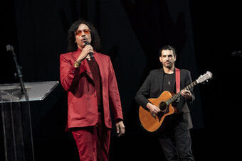 A person in a red suit sings in front of a person playing guitar.