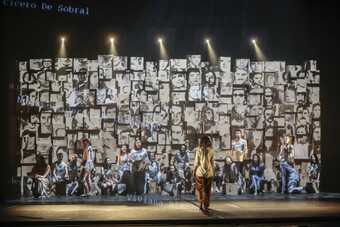 Actors stand in front of a collage of faces along the back wall of a theatre