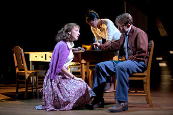 Three actors sit around a table onstage.