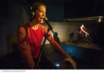 An actor onstage rides a stationary bike while speaking into a microphone.