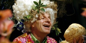 An actor wearing a large curly wig smiles at a prop in their hand.