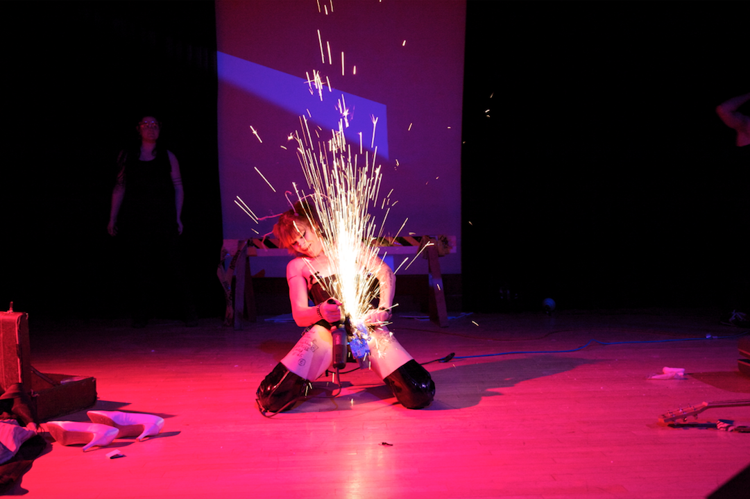 performer kneels onstage while sparks fly between their legs