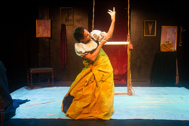 An actor in a yellow period dress gestures on stage.