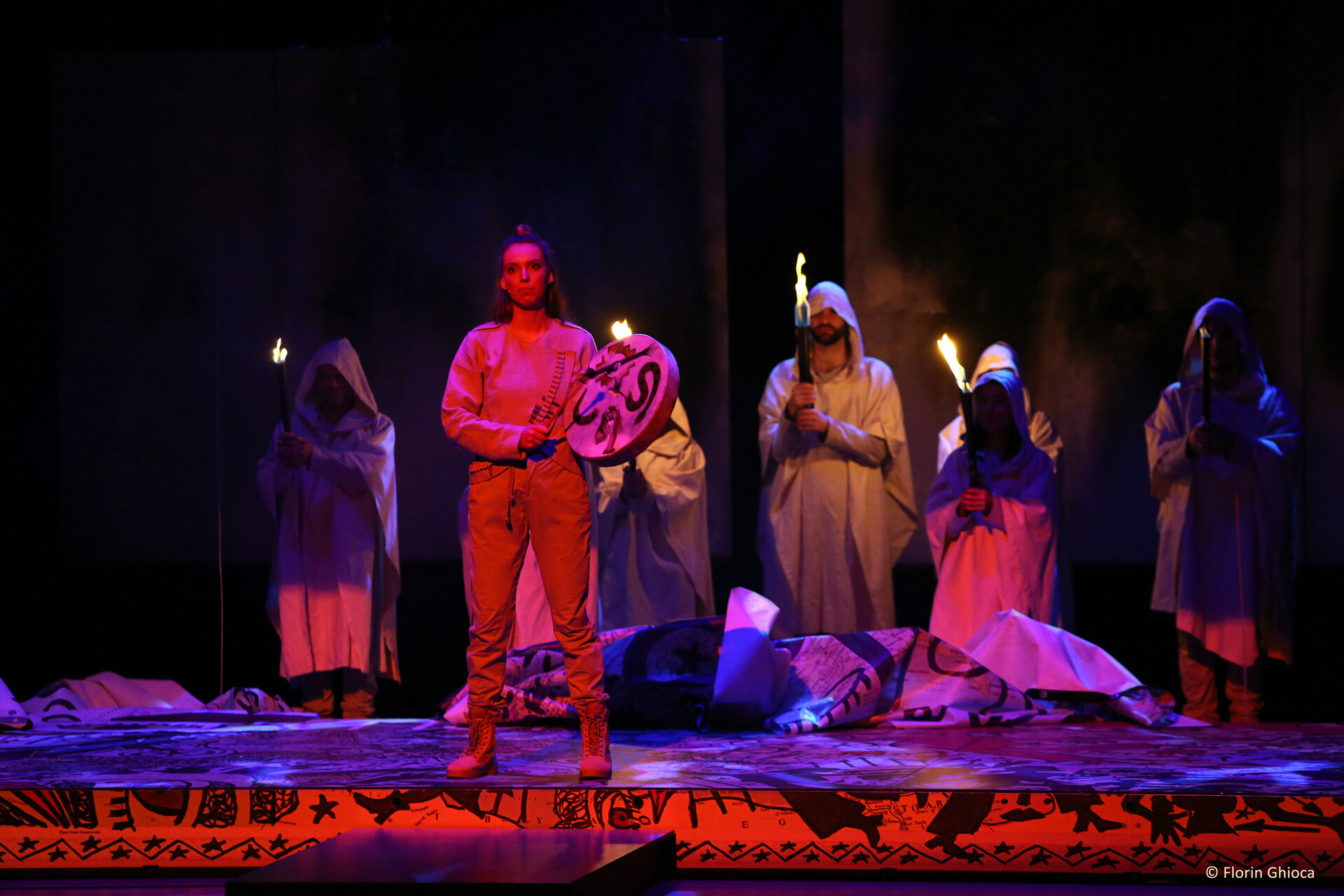 performers onstage in hoods holding torches, one person standing and holding a drum