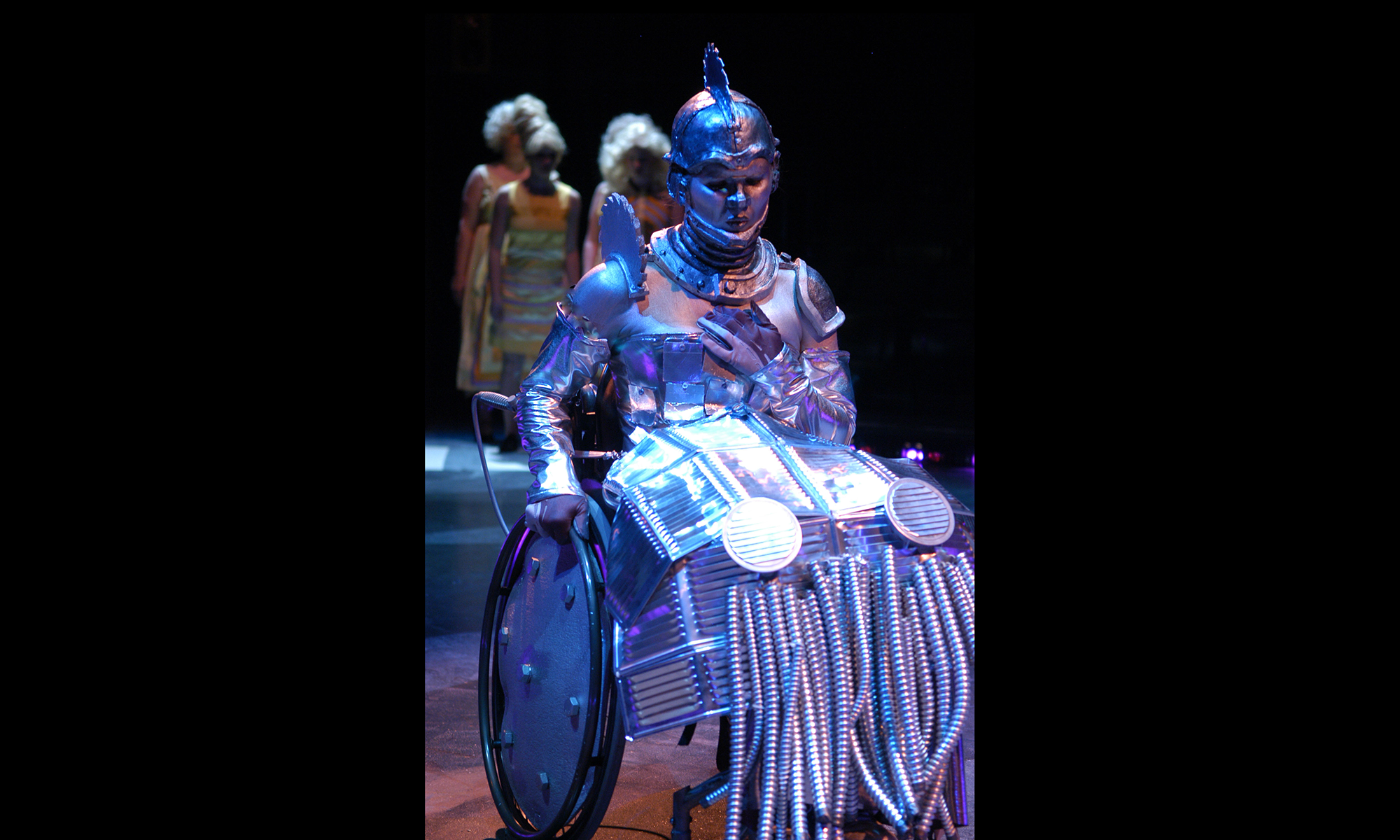 wheelchair user in dramatic lighting in tin metal costume from head to wheel, saw blade on top of head and shoulders, metal-plated corset and metal skirt
