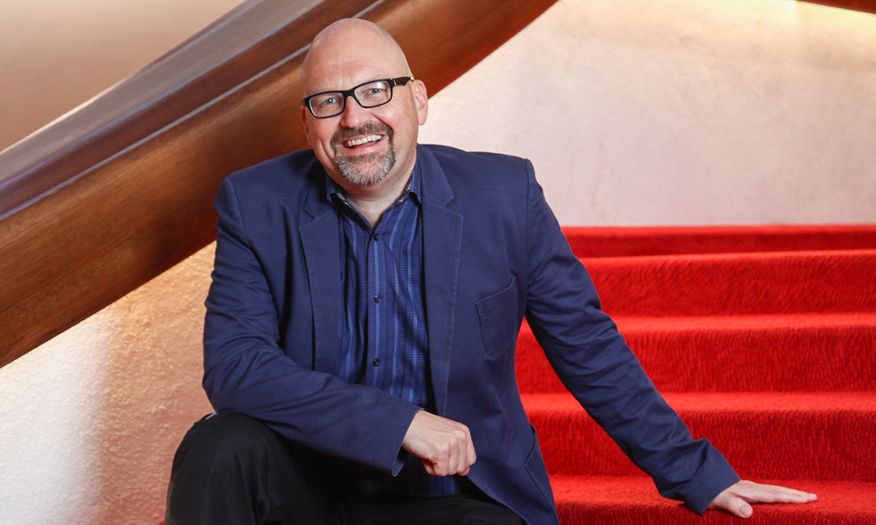a bald man with glasses and a beard in a navy suit sitting on red stairs