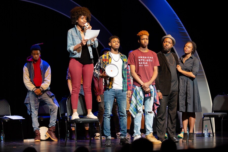 a group of Black youth and adults stand onstage while one youth speaks into a megaphone