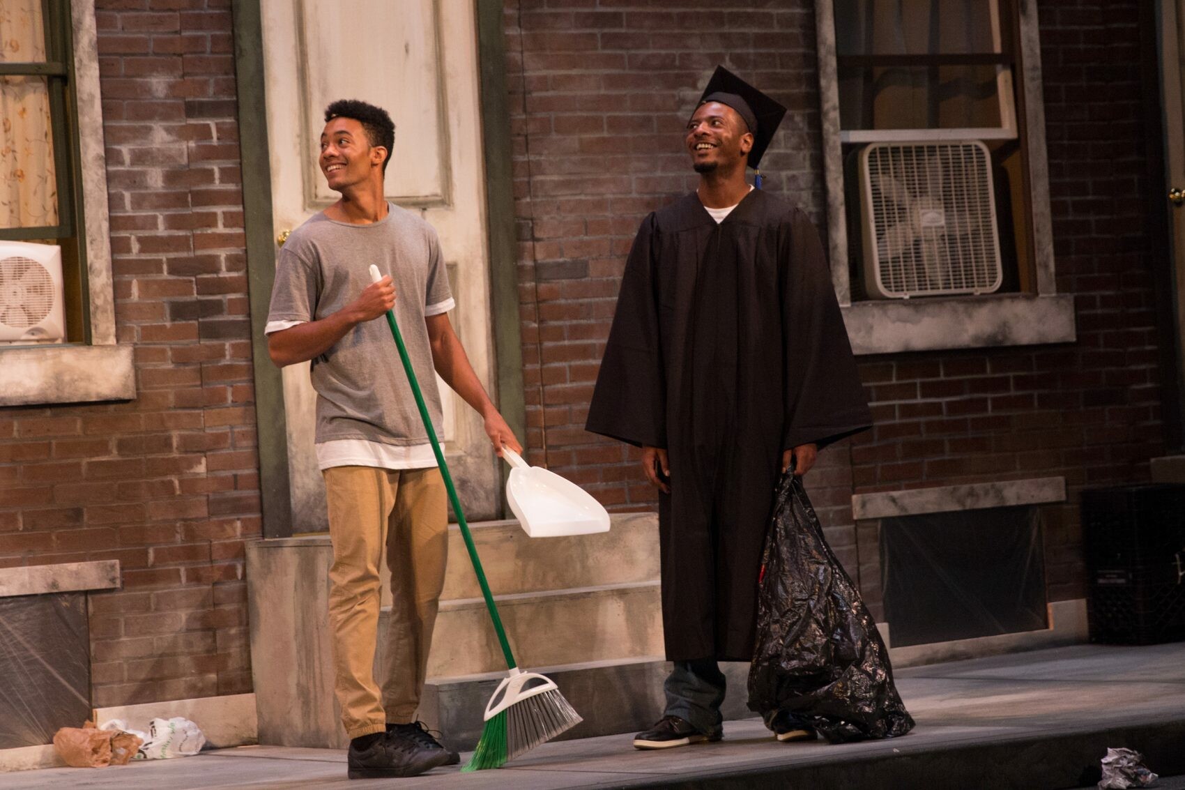 two actors on a porch, one holding a broom, and the other in a cap and gown holding a trash bag.