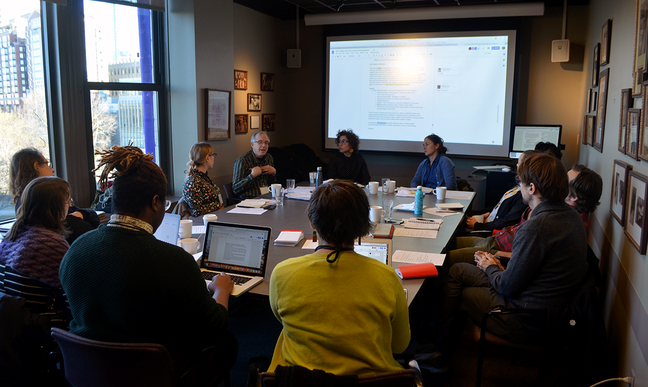 A group of theatre artists sitting around a conference table with a projector screen in the background.