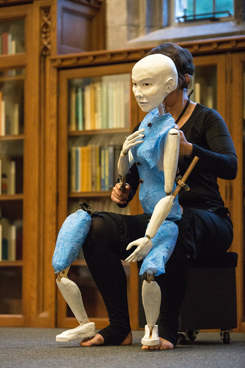 Daylight lighting. The puppeteer, dressed in black, is sitting on a short black cube on a grey stage in front of bookshelves. The puppet, with white body and light blue costume, is sitting on the puppeteers knee. The puppet head, bald and painted off-white, is blocking the puppeteers face. The puppet's right leg is separate from the rest of its body, connected to the right leg and toes of the puppeteer. There is a large space between the two puppet legs. The gesture of the right hand of the puppet is raised