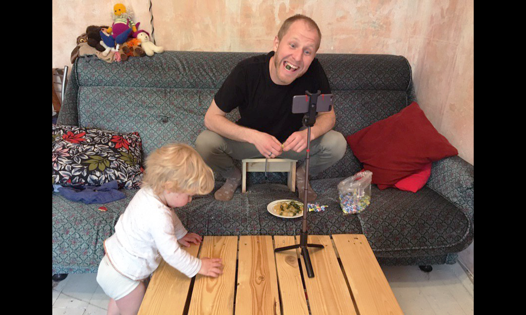 a person seated on a couch recording themselves on a phone situated in a stand on a coffee table. their child is to their right with their hands on the table