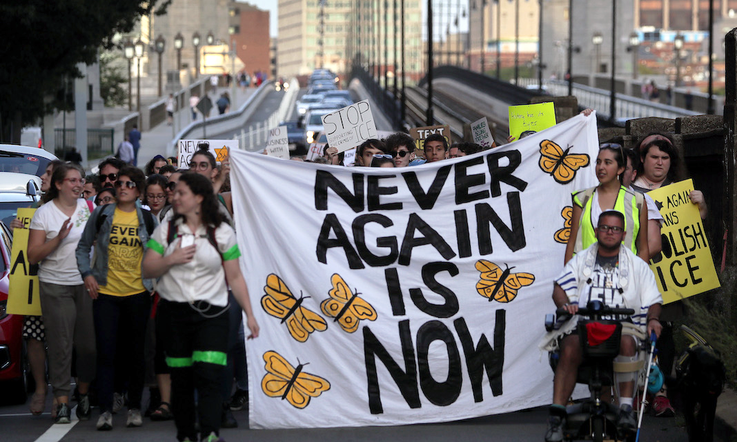 a group marching outside with a banner that reads "NEVER AGAIN IS NOW"