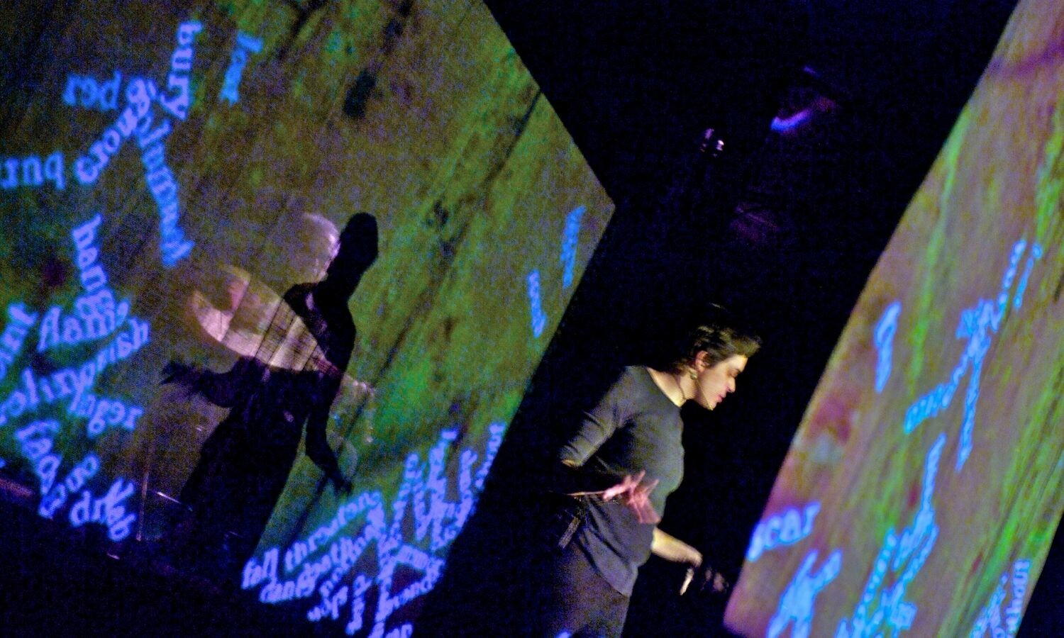 Andrea stands facing one screen with projected words on it with another behind her. Behind the second screen stands a man playing the oud. 