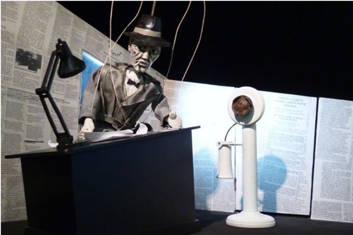 Puppet is standing behind a desk made to scale with tiny lamp and white phone. Newspaper clippings compose the backdrop. 