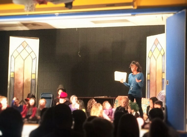 An image of a person standing on stage in a school auditorium surrounded by elementary school students. 