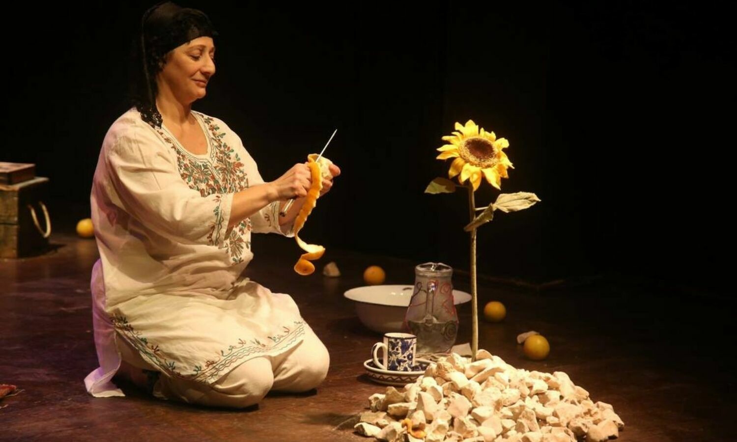 A woman dressed in white kneeling on the floor and cutting off the peel of an orange. She sits in front of one sunflower.