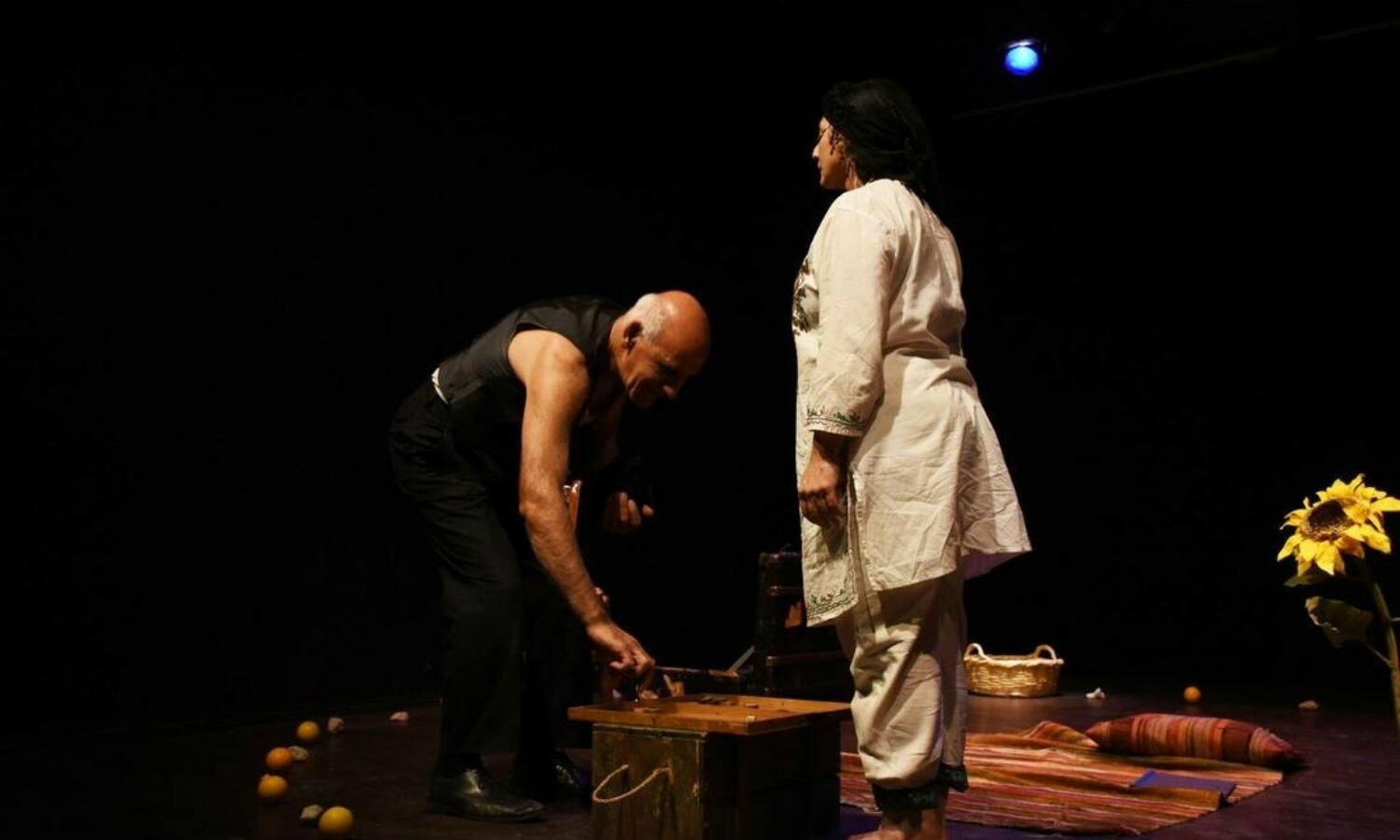 A man on the left bending down to pick things off a stool and a woman on the right standing up straight and looking down at him.