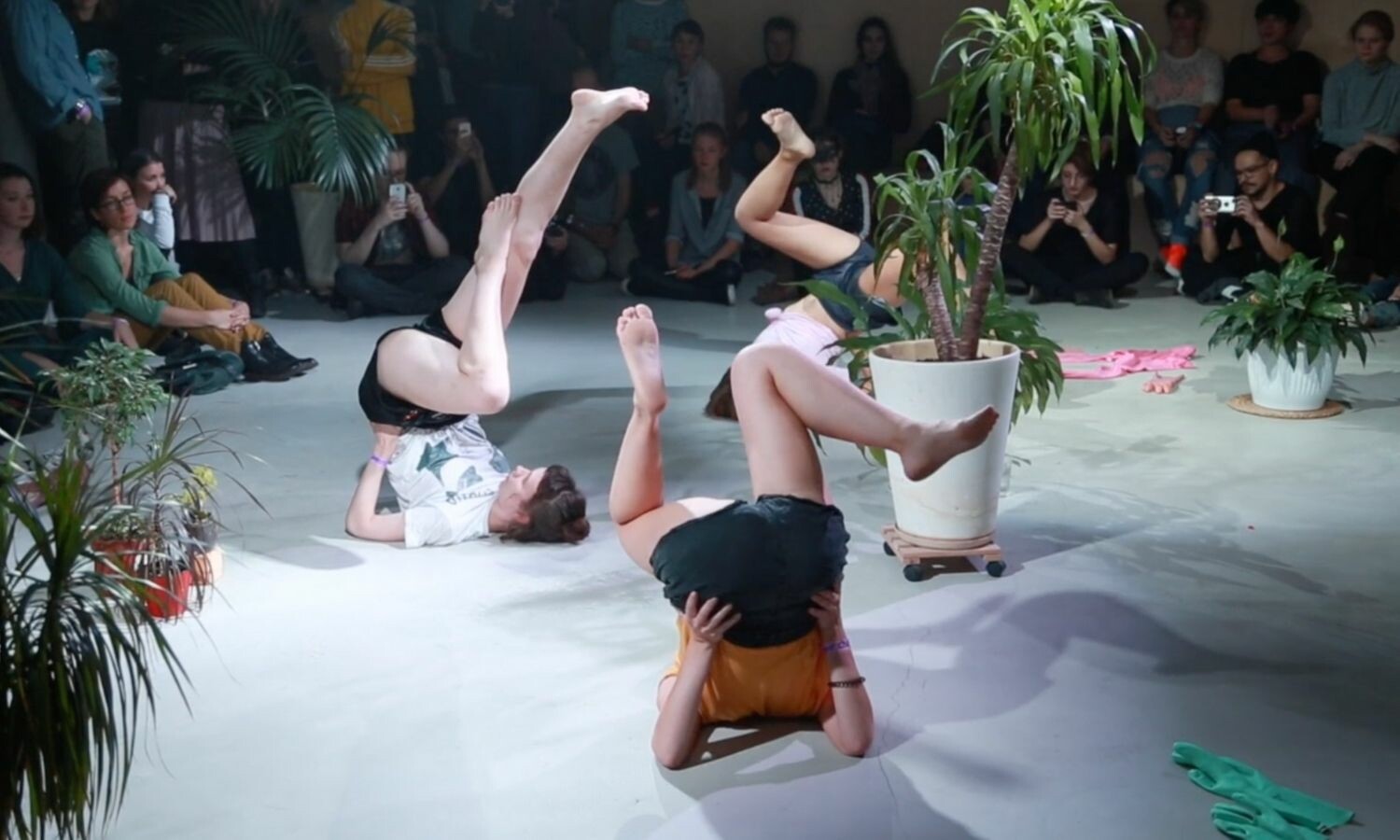 Three actors with their heads and shoulders on the ground and legs up. They're surrounded by the audience and some plants.
