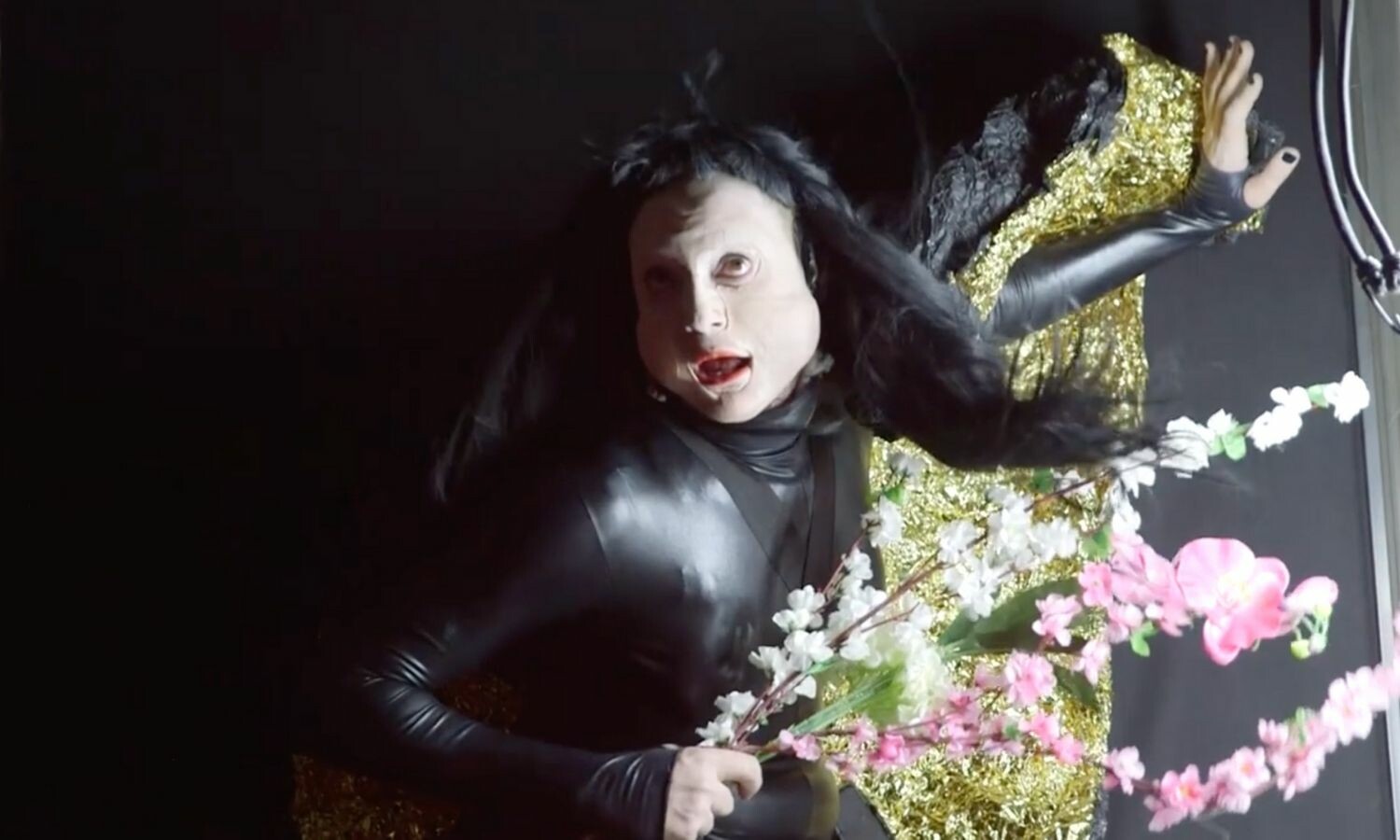 An actor with long black hair in a leather bodysuit, wearing black nail polish and holding flowers.