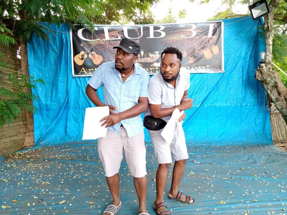 Image of two men standing in front of a banner rehearsing outdoors. They are huddled close to each other and one is pointing into the distance, while the other looks in the same direction. 