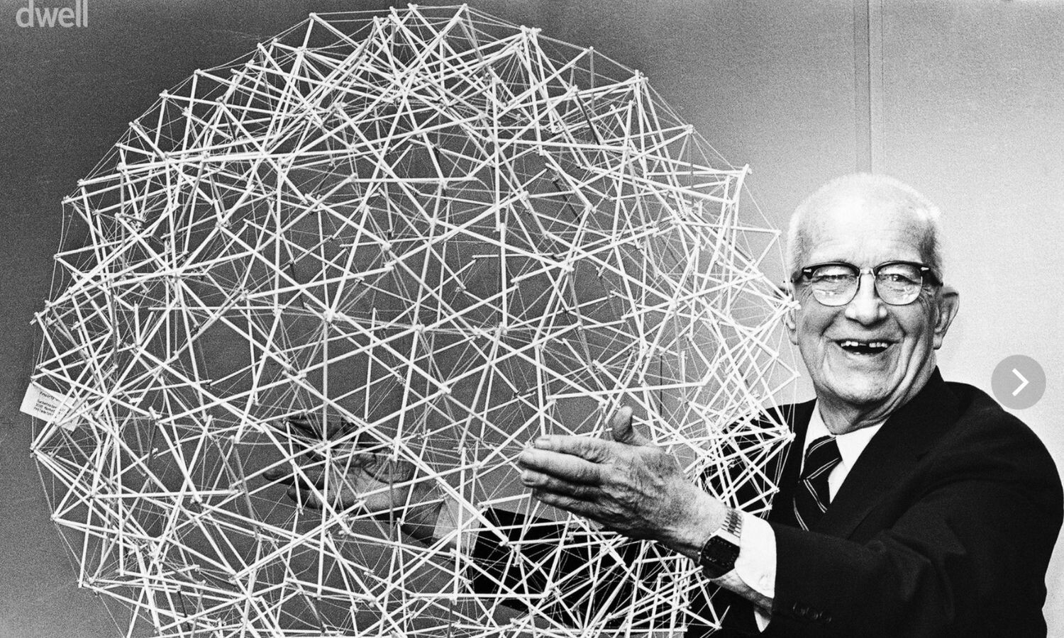 Buckminster Fuller smiling and holding a sphere made of sticks connected by string.