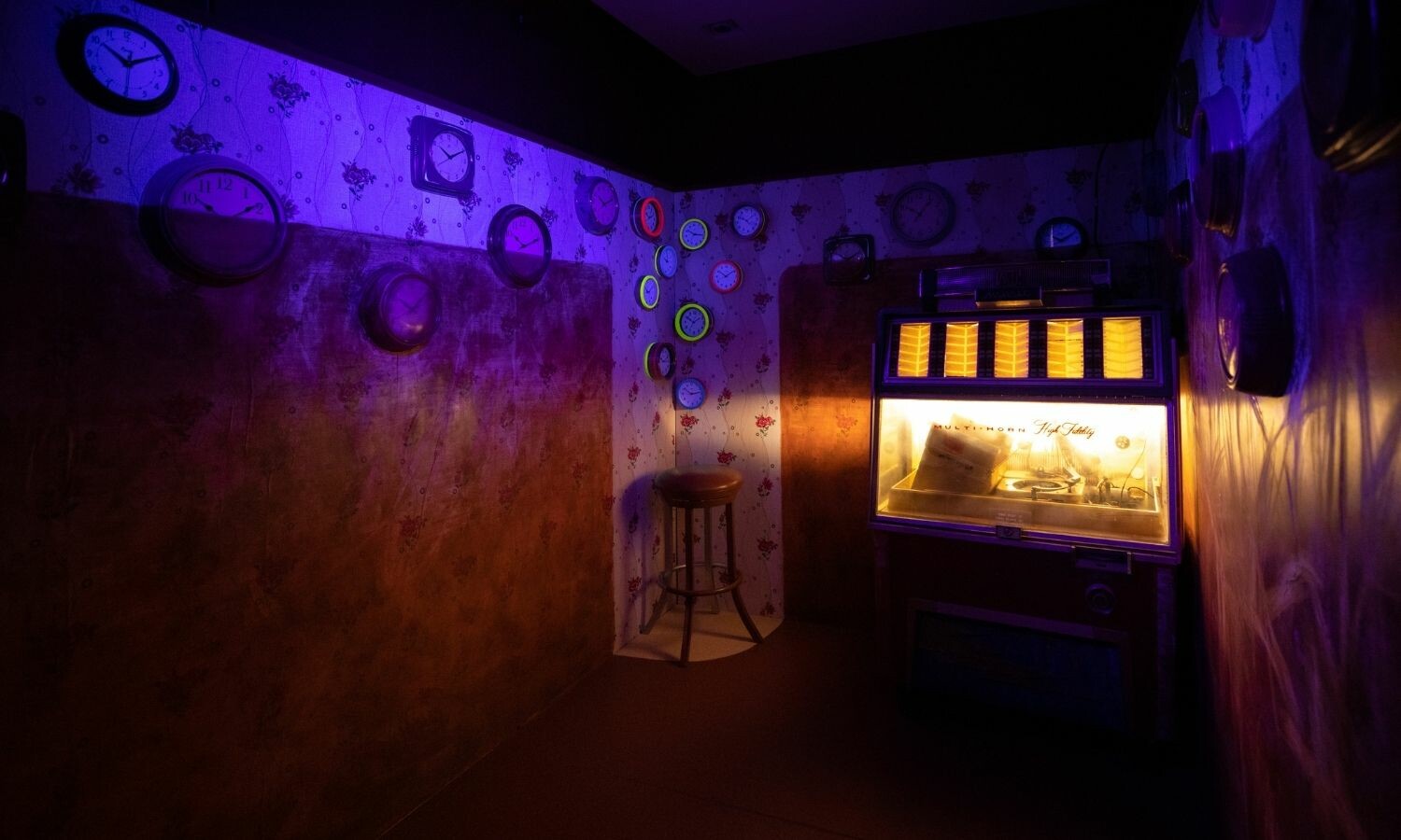 A dimly lit room with purple lighting, several clocks on the walls, a stool, and a jukebox.
