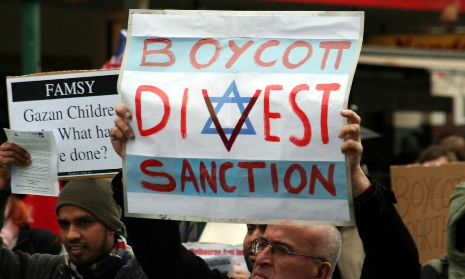 A man holding an Israeli flag with the words "Boycott, Divest, Sanction" on each of the three stripes. The 'v' in "Divest" goes right over the Star of David.
