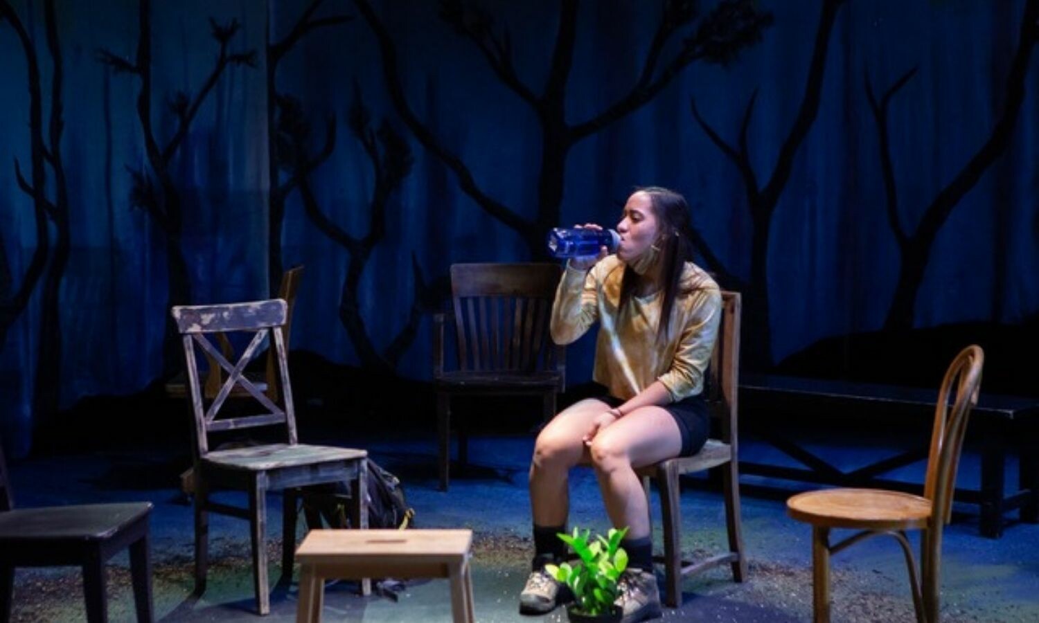 A woman drinking water and sitting down on a chair surrounded by other chairs and onstage "woods".